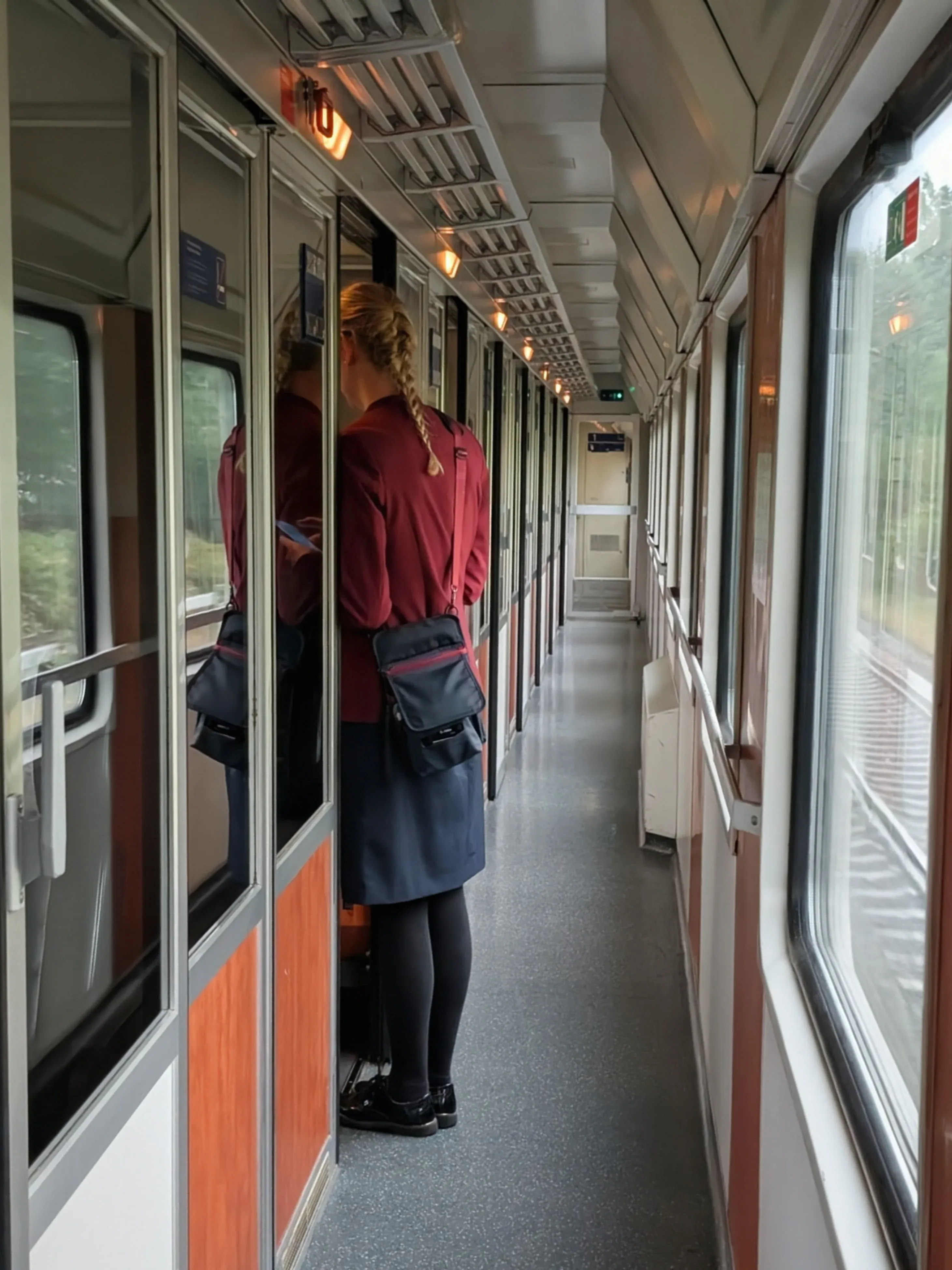 A conductor in a red uniform checks tickets at a compartment door in the side corridor of an older compartment coach near Bad Endorf, Germany.