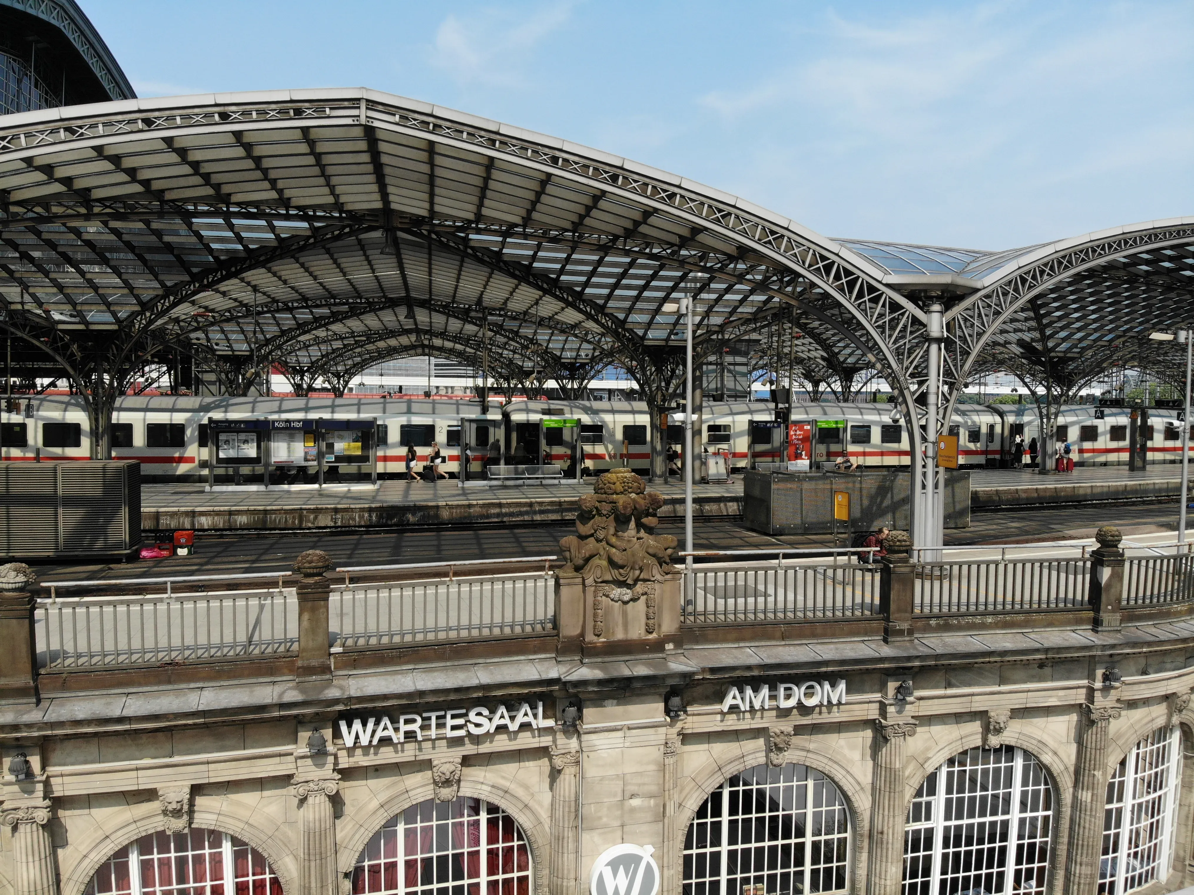 Historic stone facade of the Wartesaal am Dom overlooking the arched iron-and-glass train shed of Köln Hauptbahnhof with a German ICE train at the platform.