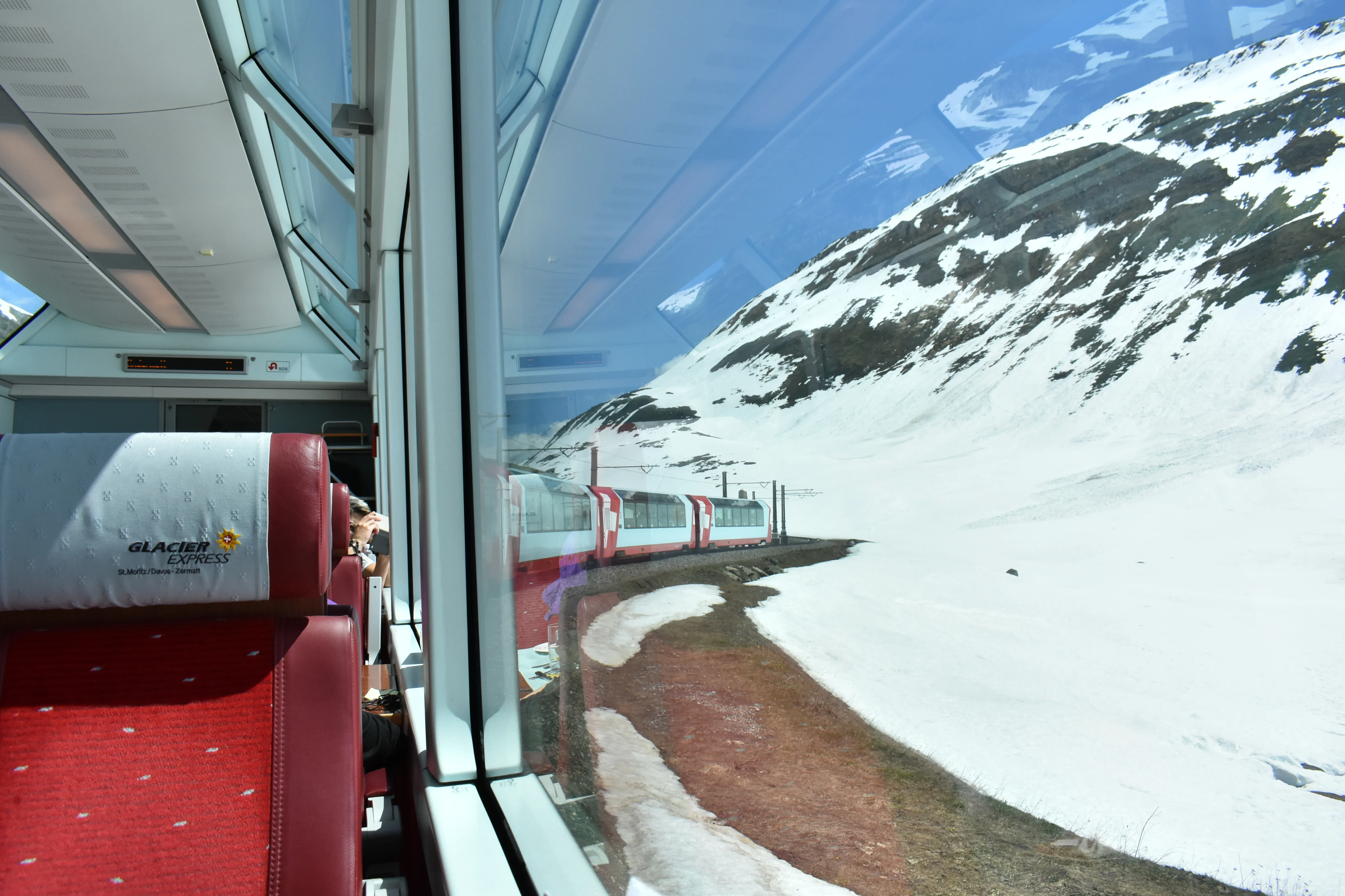Interior view of a Glacier Express panoramic train carriage looking out at deep alpine snowbanks while crossing the Oberalp Pass in Switzerland.