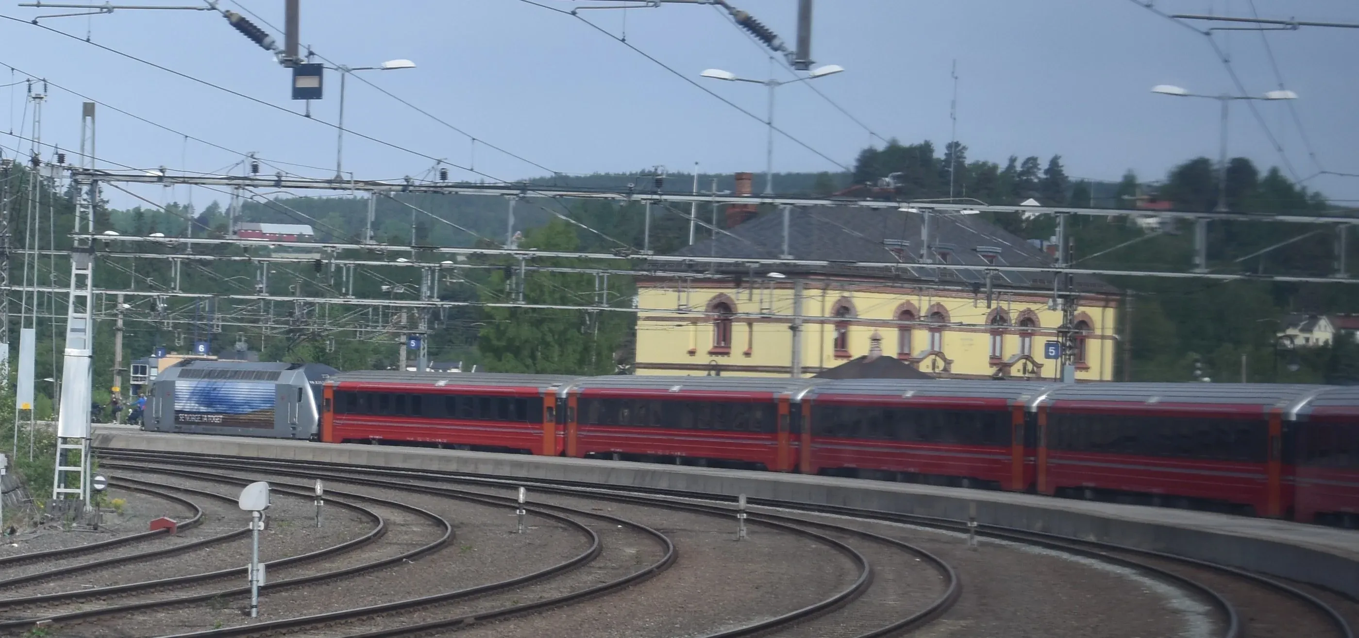 A Norwegian NSB electric locomotive and red passenger train carriages on curved railway tracks at Hønefoss station in Norway, with a yellow historic station building in the background.