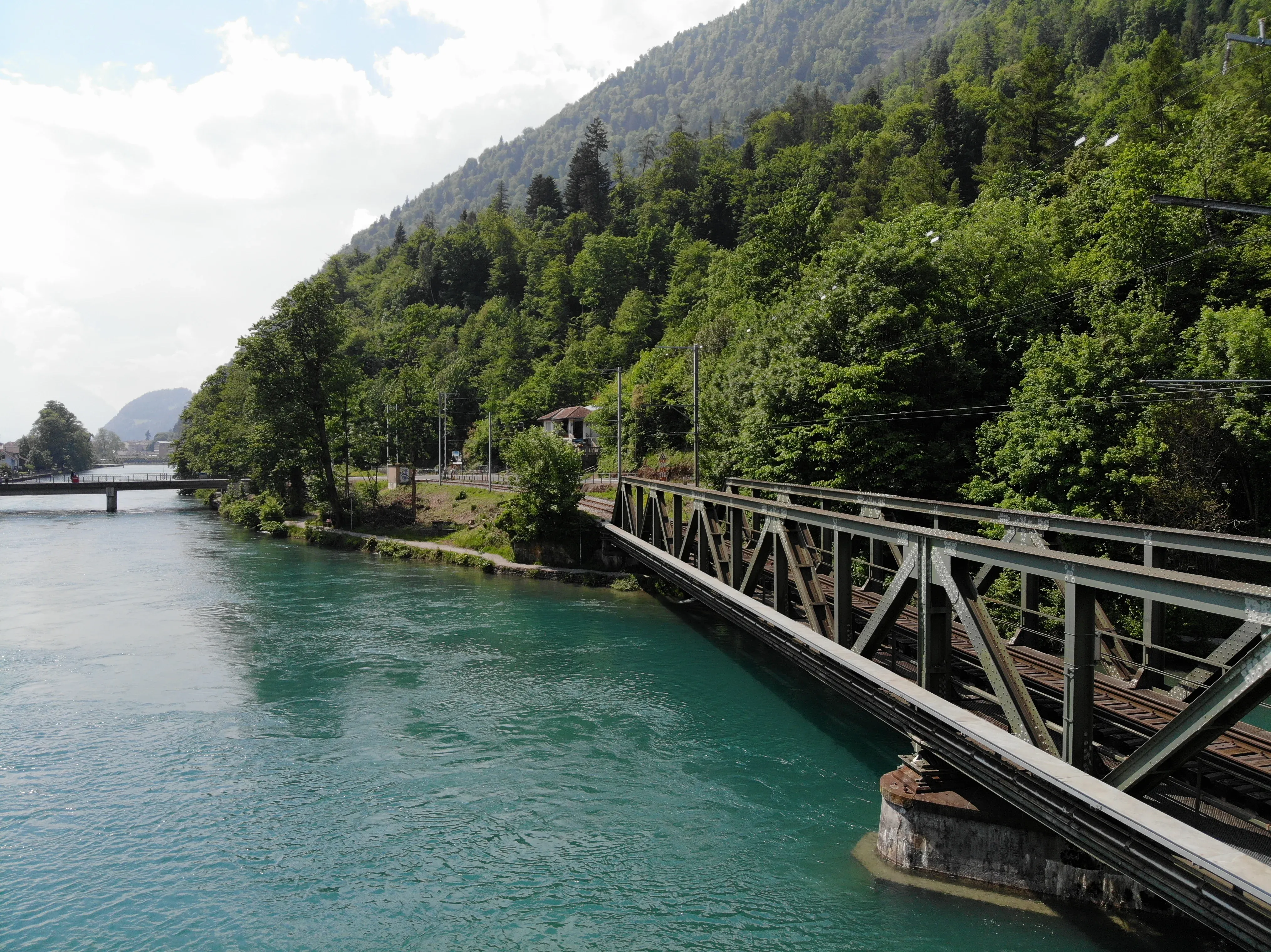 Green steel railway truss bridge over the turquoise Aare river surrounded by lush forested mountains in Interlaken, Switzerland.