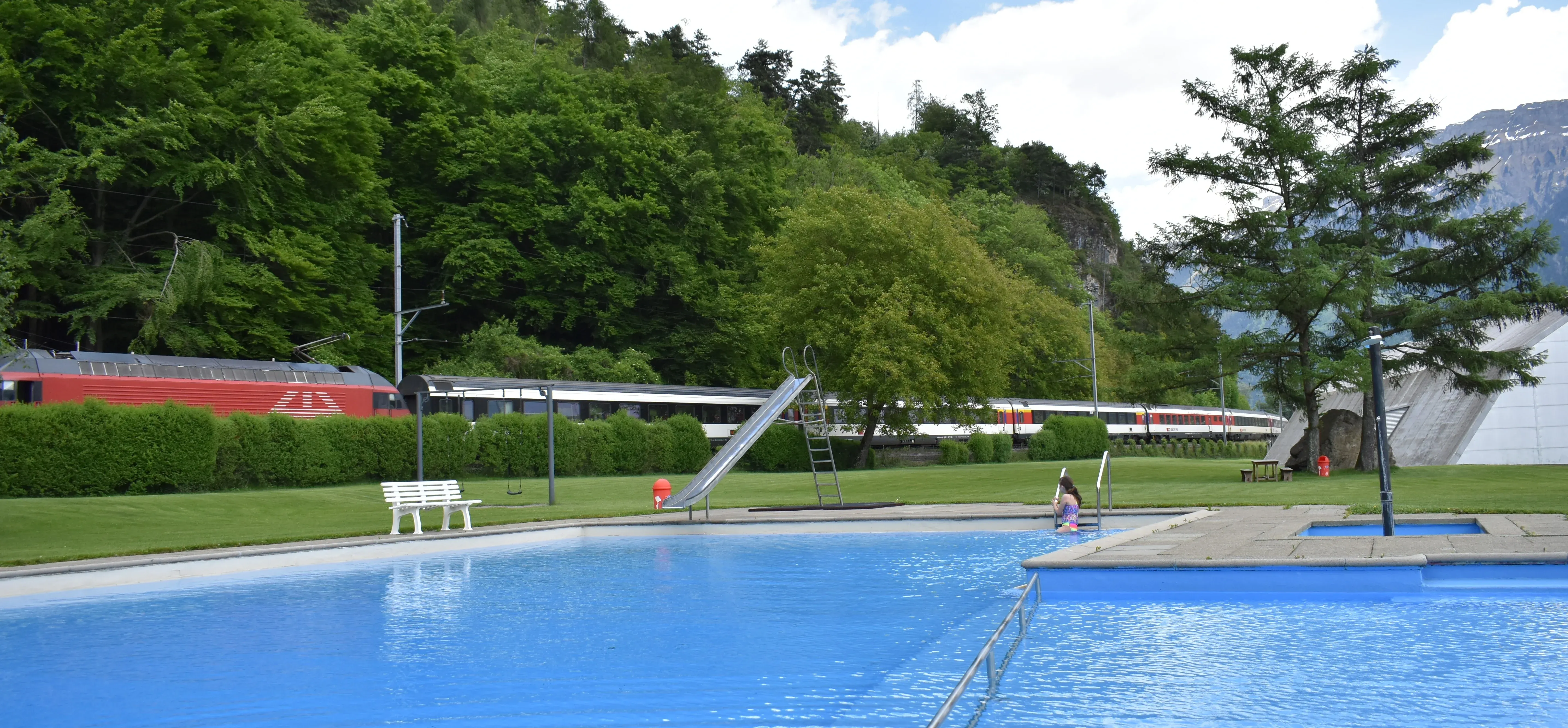A red Swiss SBB electric locomotive and passenger train speed along the railway past the outdoor pool and lawn of Bödelibad in Interlaken, Switzerland.