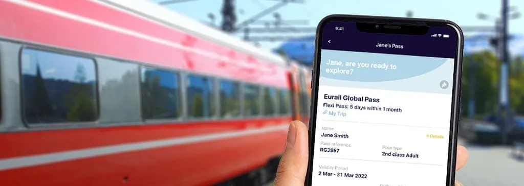 A traveler holding a smartphone displaying an Interrail mobile pass barcode with a passenger train on the platform in the background.