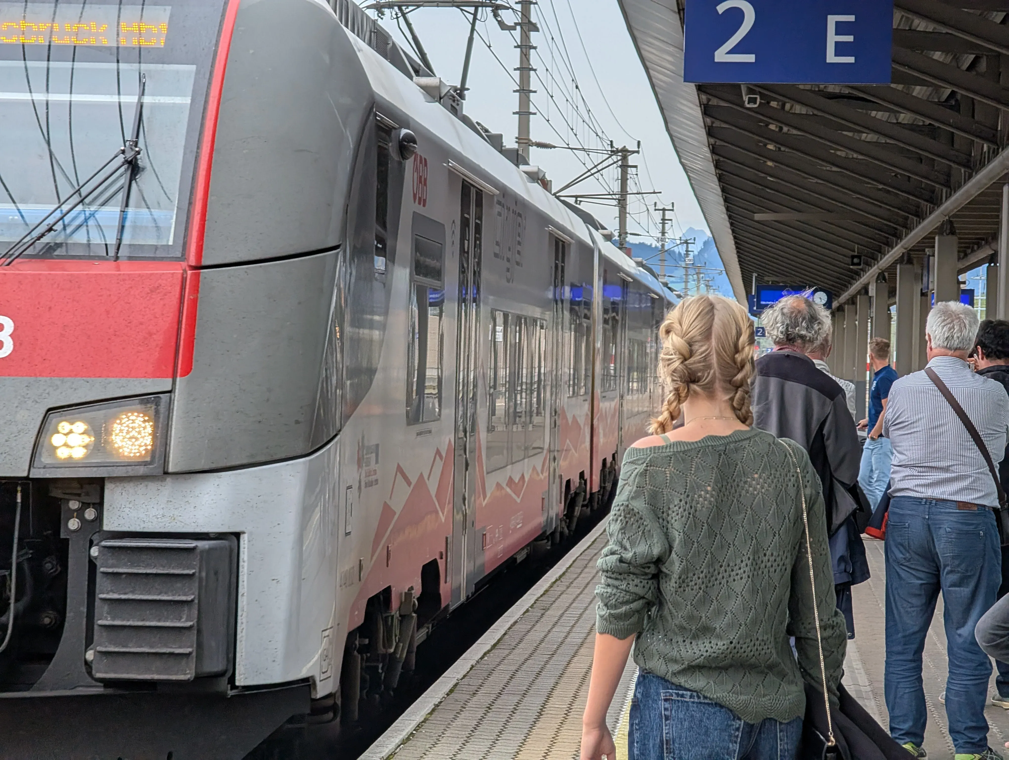 Passengers waiting on a covered platform marked 2 E beside a red and silver ÖBB regional train at Jenbach station in Tyrol, Austria.