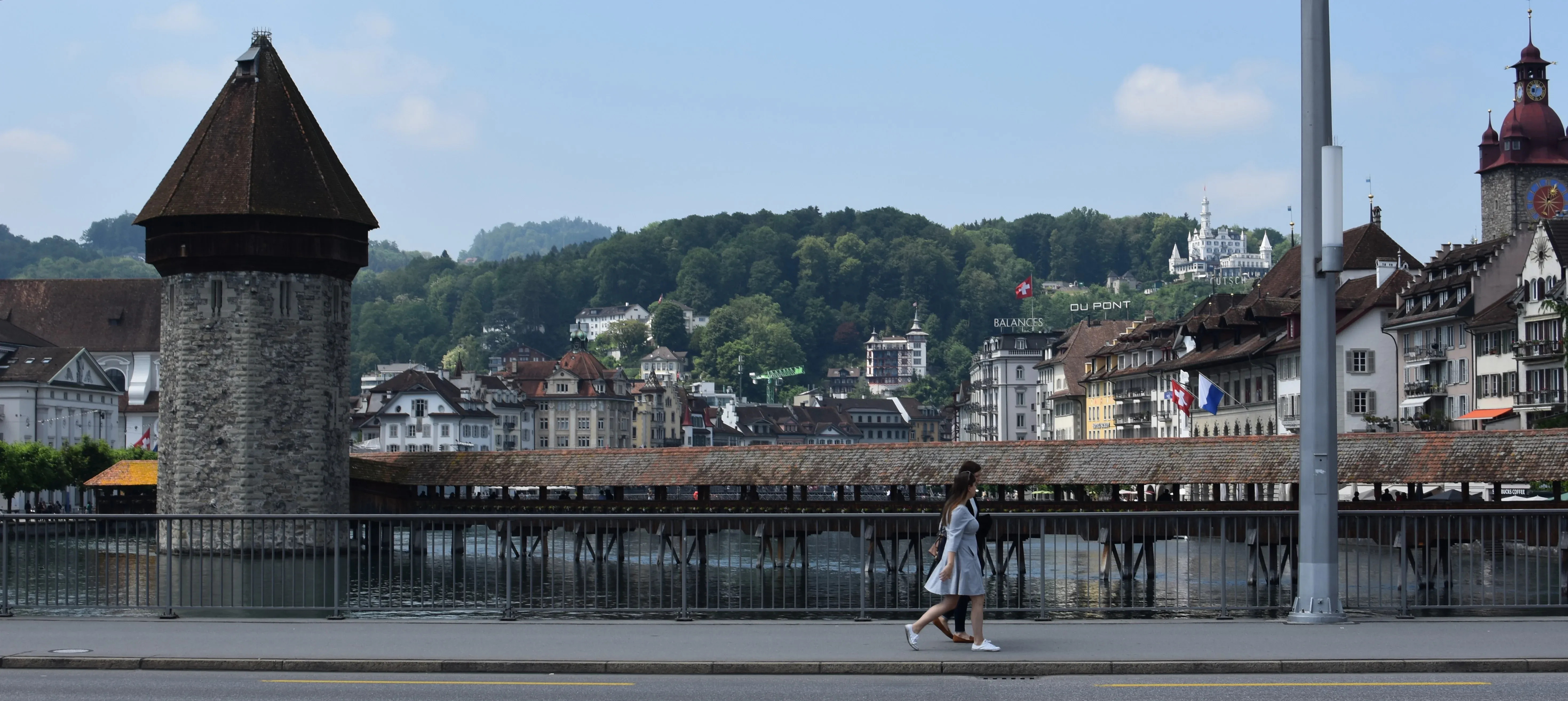 Pedestrians walking along the waterfront past the historic wooden Chapel Bridge and stone Water Tower on the Reuss river in Lucerne, Switzerland.