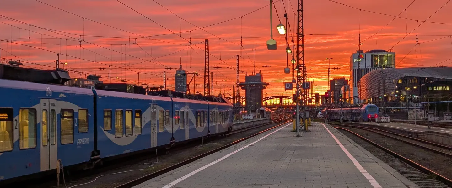 Looking across the open-air railway platforms and tracks of München Hauptbahnhof toward a vibrant orange sunset in Munich, Germany.