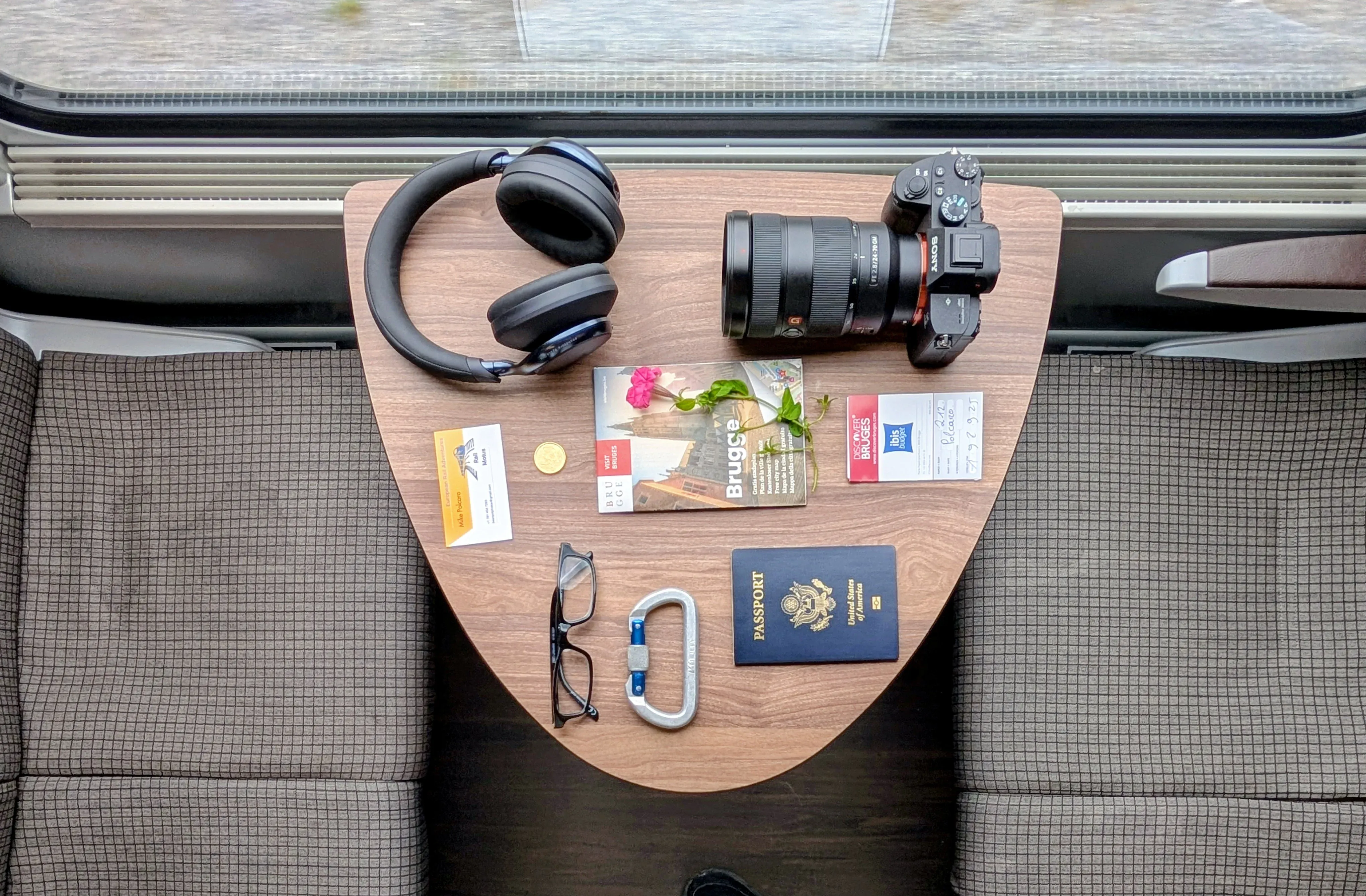 Top-down flatlay of travel essentials including a passport, mirrorless camera, eyeglasses, and headphones laid out on a wooden train tray table in Belgium.