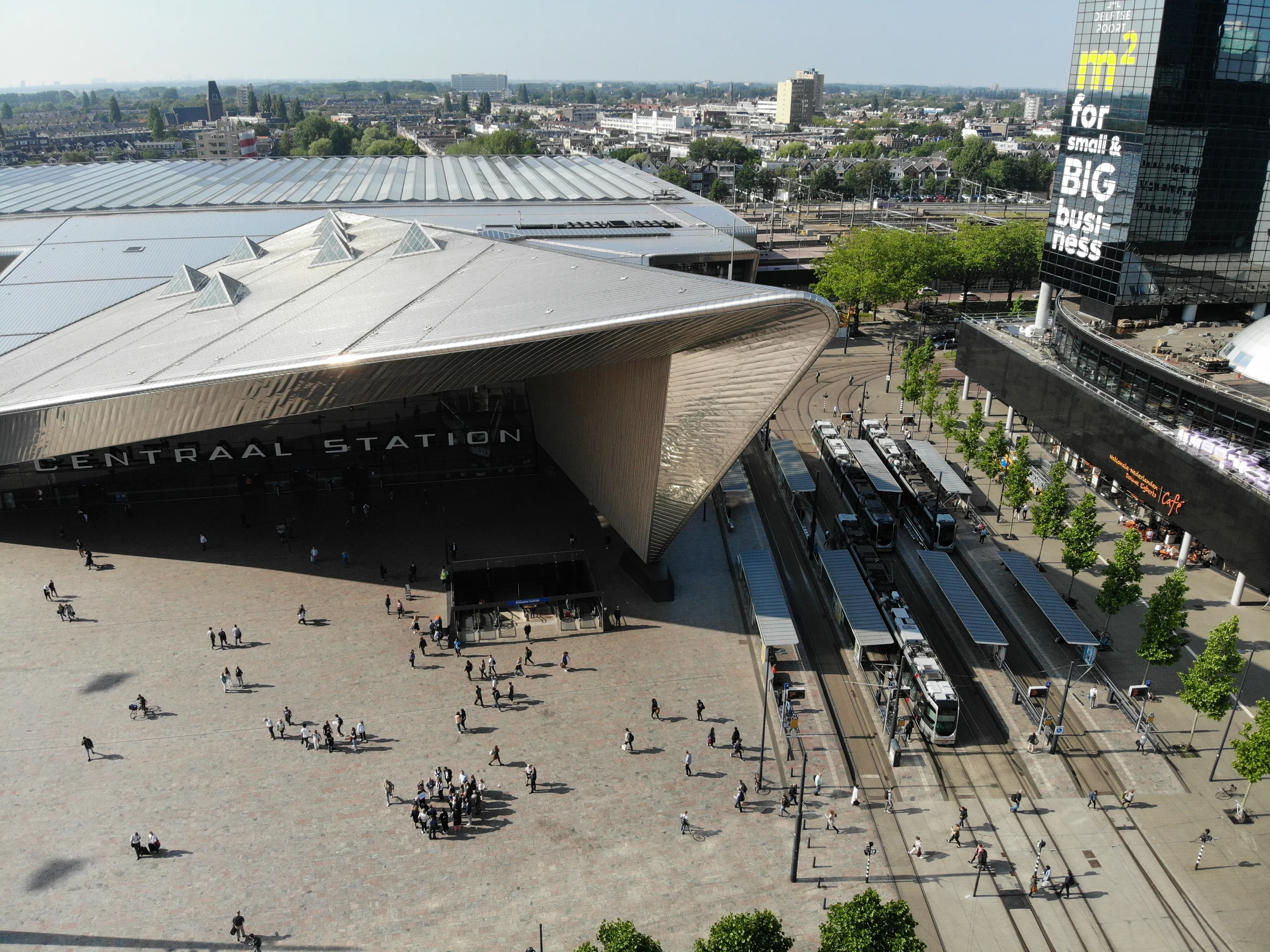 Aerial perspective of the dramatic angular stainless-steel canopy of Rotterdam Centraal station and the bustling pedestrian forecourt with trams in the Netherlands.