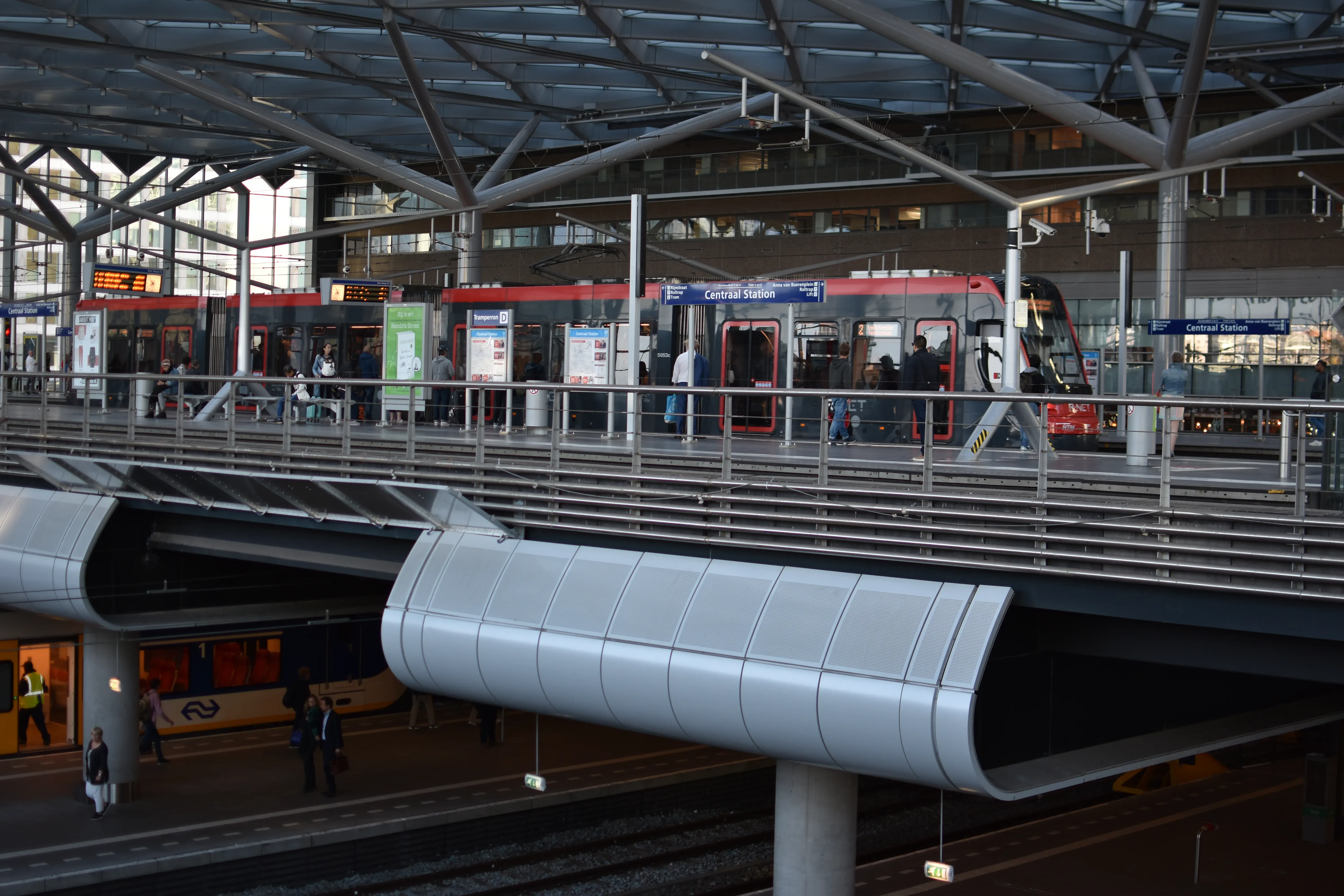 Elevated tram platform crossing over train tracks inside the glass-roofed The Hague Central Station, with a gray and red HTM tram waiting for passengers.