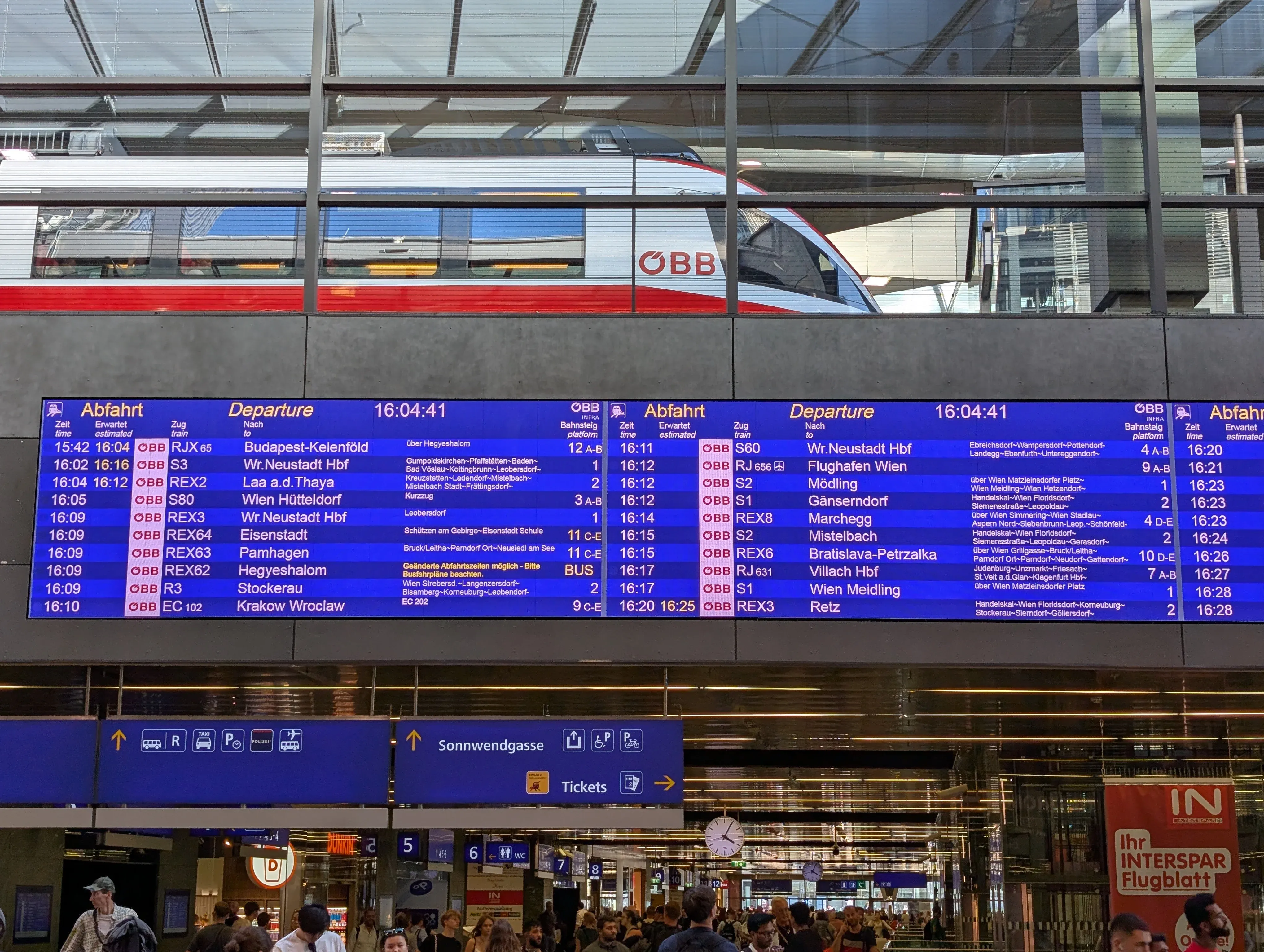 The towering blue digital departure board and bustling concourse of Vienna Central Station (Wien Hauptbahnhof) showing international Railjet and regional train departures.